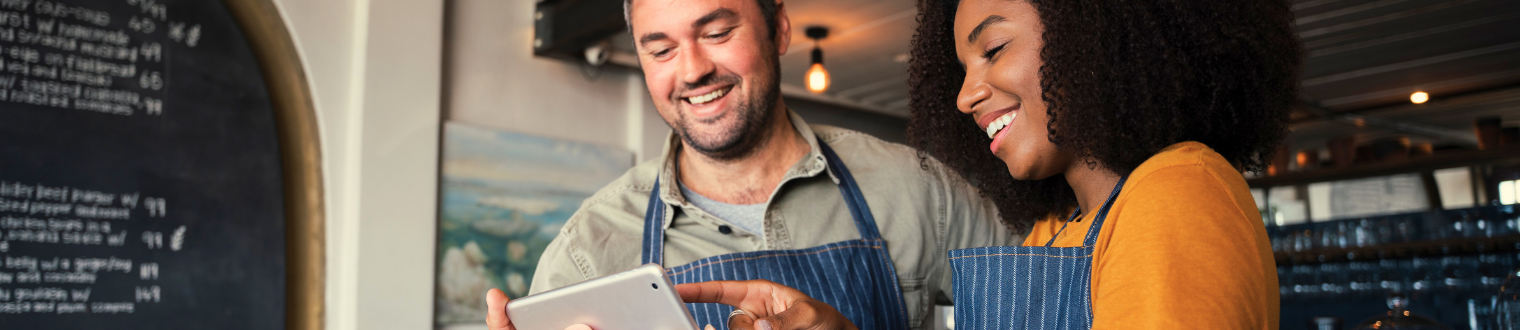 Male and Female small business owners in coffee shop smiling and looking at a tablet.